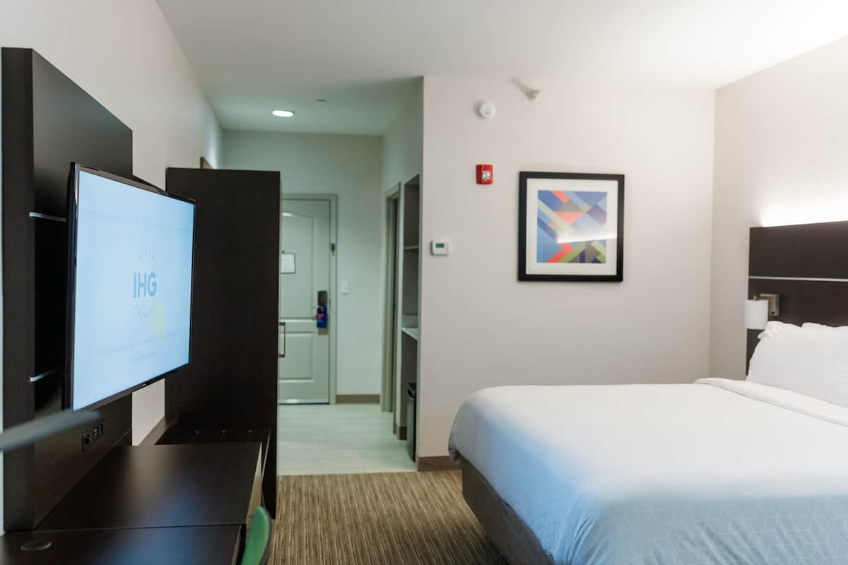 A hotel room with a bed, a TV, a framed abstract art piece, and a door leading to a hallway.