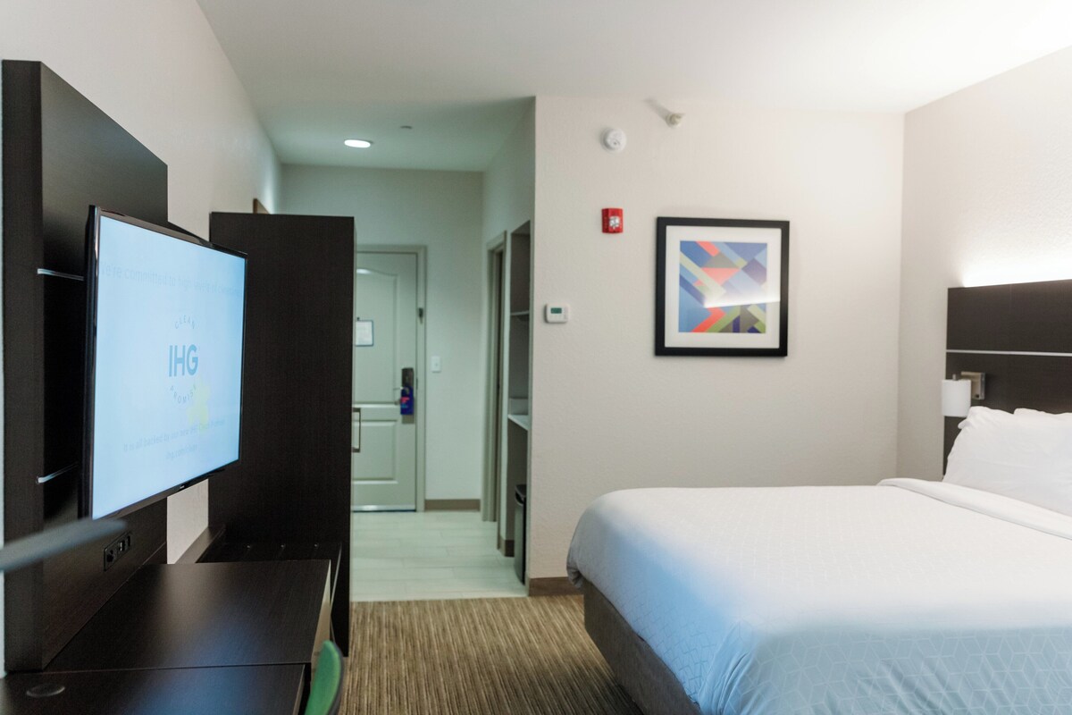A hotel room with a bed, a TV, a framed abstract art piece, and a door leading to a hallway.