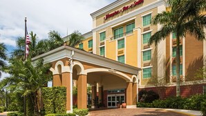 Edificio de Hampton Inn & Suites con entrada despejada, palmeras y una bandera estadounidense.