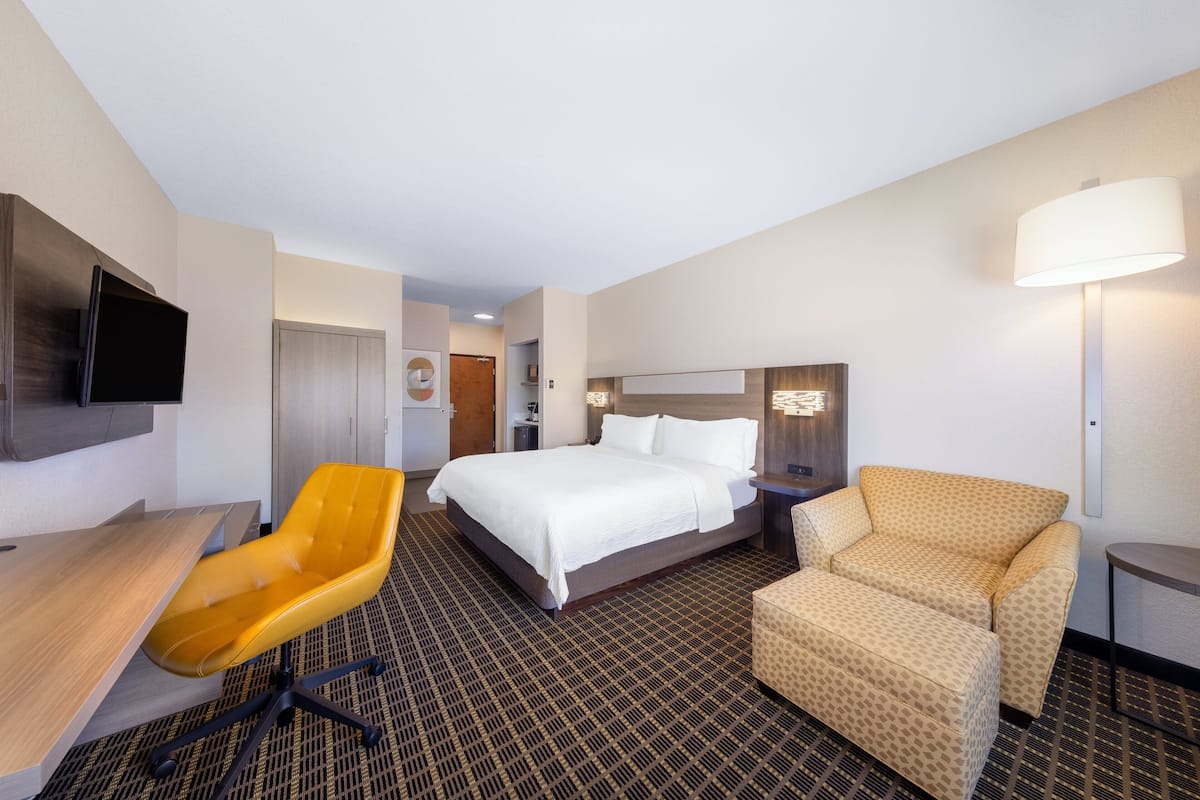 A hotel room with a bed, a desk, a yellow chair, a patterned armchair, a TV mounted on the wall, and a lamp.