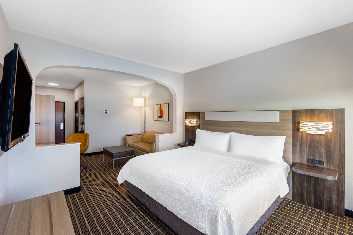 A modern hotel room with a large bed, a television, a seating area with a chair, and a small table.