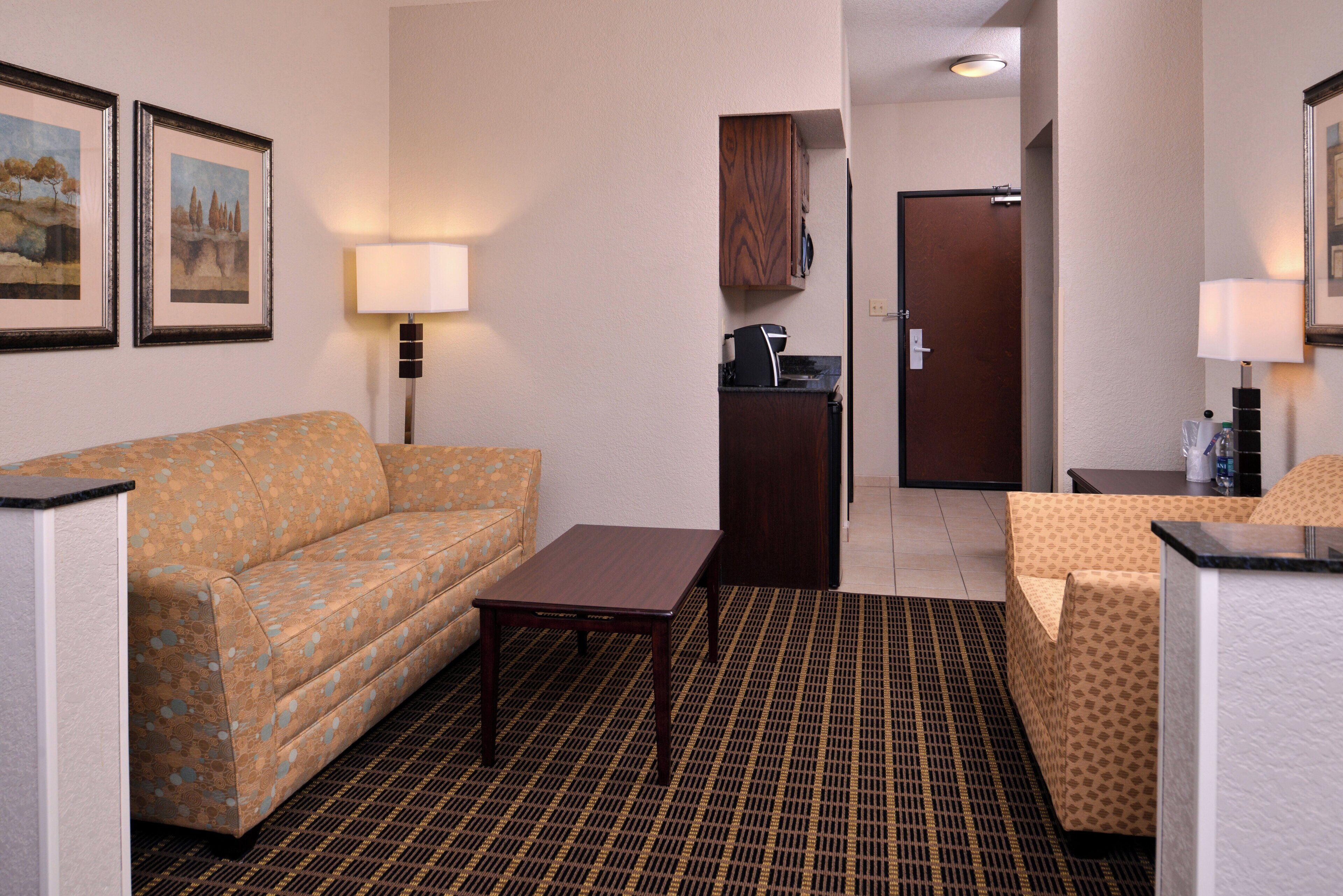 A hotel room with a sofa, a small table, a kitchenette, and a door leading to another room.