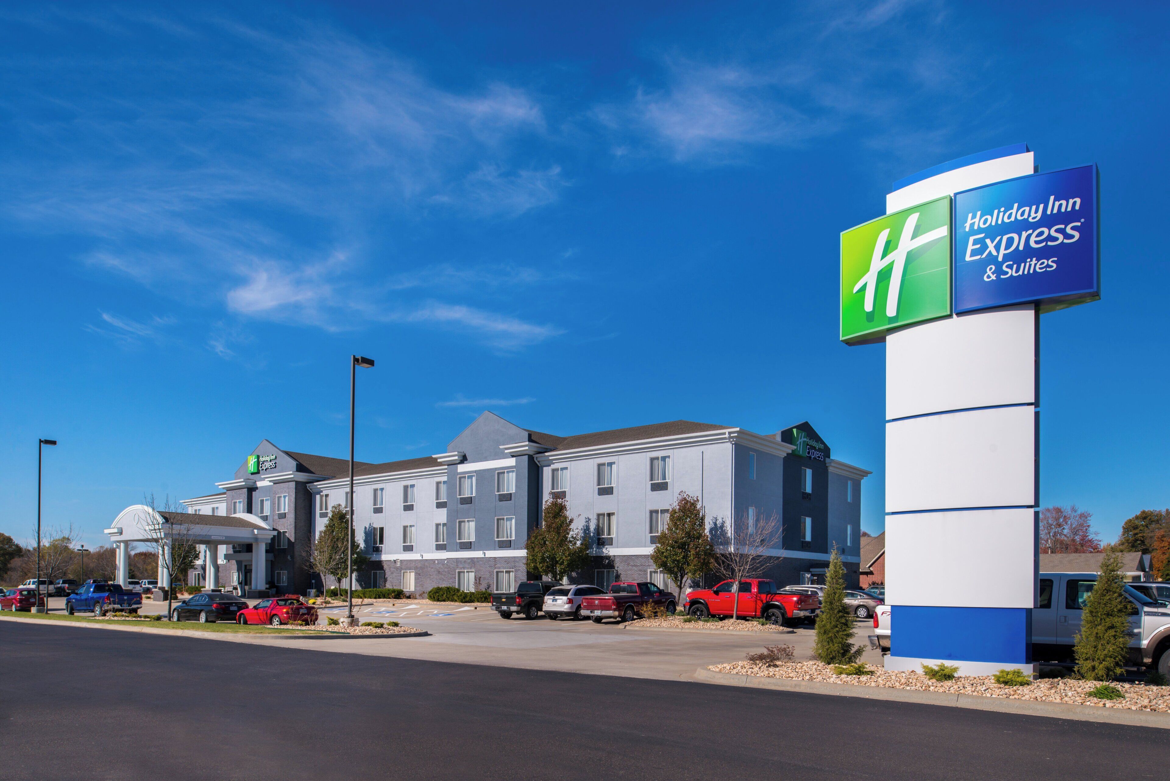 Un hotel Holiday Inn Express con diseño moderno, que destaca un letrero llamativo, un estacionamiento con varios vehículos y un cielo azul despejado.