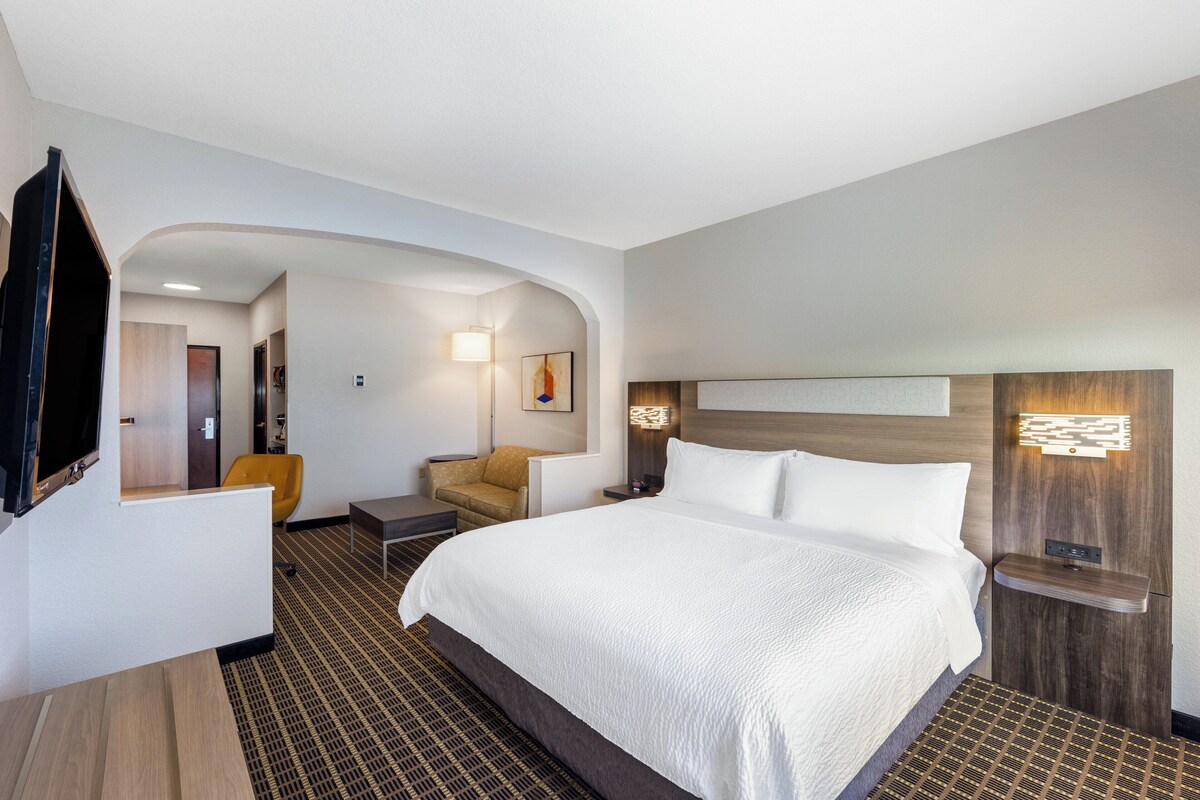 A modern hotel room with a large bed, a television, a seating area with a chair, and a small table.
