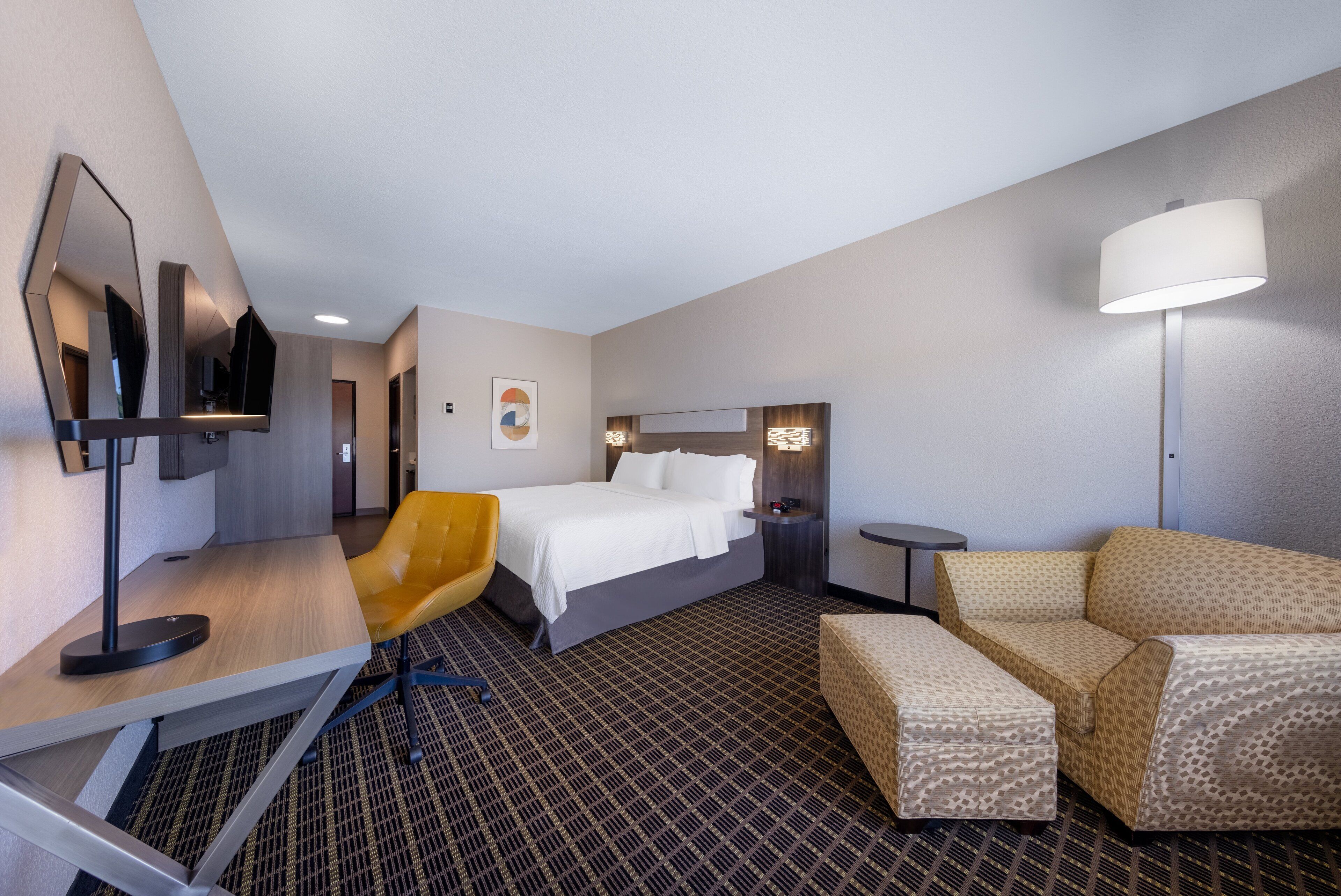 A hotel room with a bed, a desk with a chair, a sofa, a lamp, and a mirror.