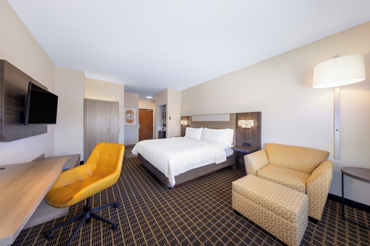 A hotel room with a bed, a desk, a yellow chair, a patterned armchair, a TV mounted on the wall, and a lamp.