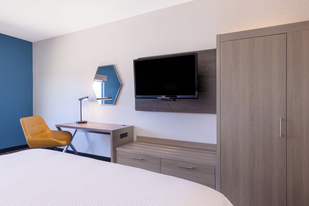 A hotel room with a bed, a desk with a chair, a flat-screen TV, a wardrobe, and a mirror on the wall.