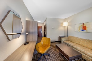 A modern hotel room with a wooden desk, a yellow armchair, a sofa, a large mirror, and a painting on the wall.