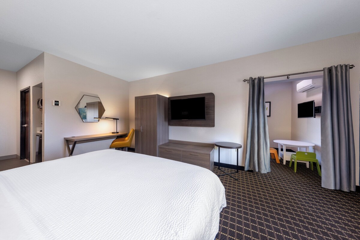 A hotel room with a large bed, a desk with a chair, a TV mounted on the wall, a small table with chairs, and a door leading to another room.