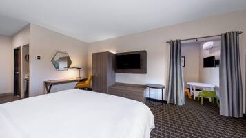 A hotel room with a large bed, a desk with a chair, a TV mounted on the wall, a small table with chairs, and a door leading to another room.