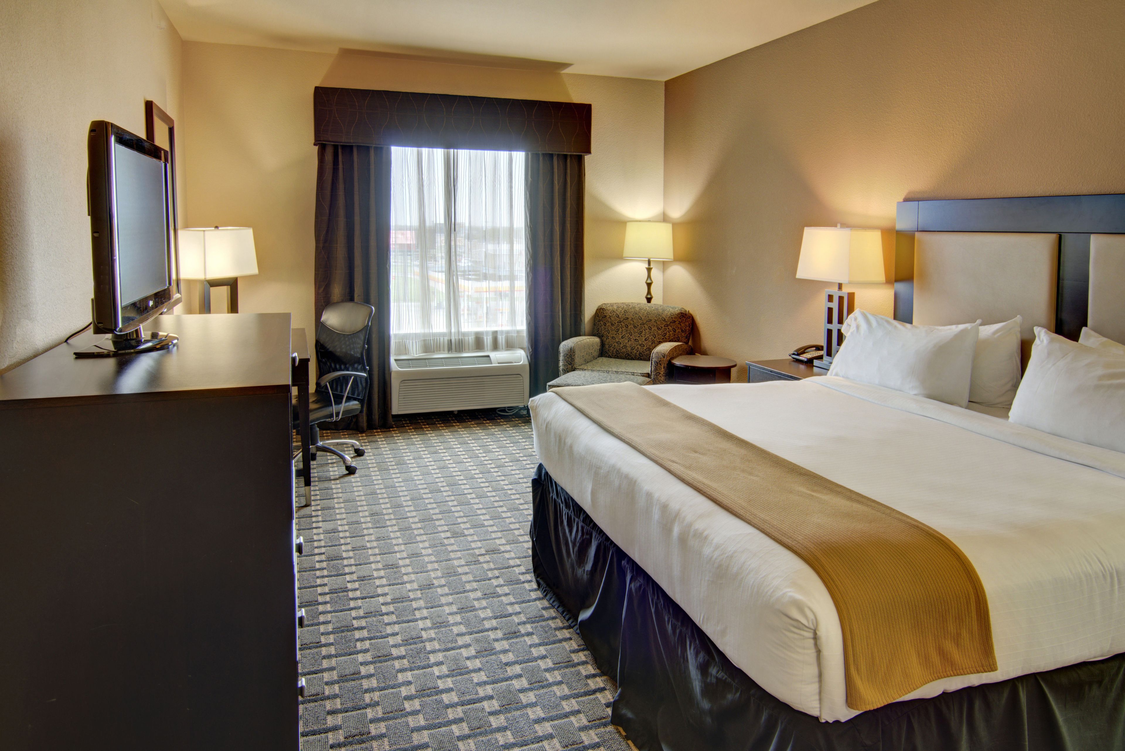 A hotel room with a large bed, a desk with a computer, a chair, a TV, and a window with curtains.