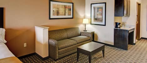 A hotel room with a bed, a sofa, a small table, a lamp, a framed picture, and a kitchenette area.