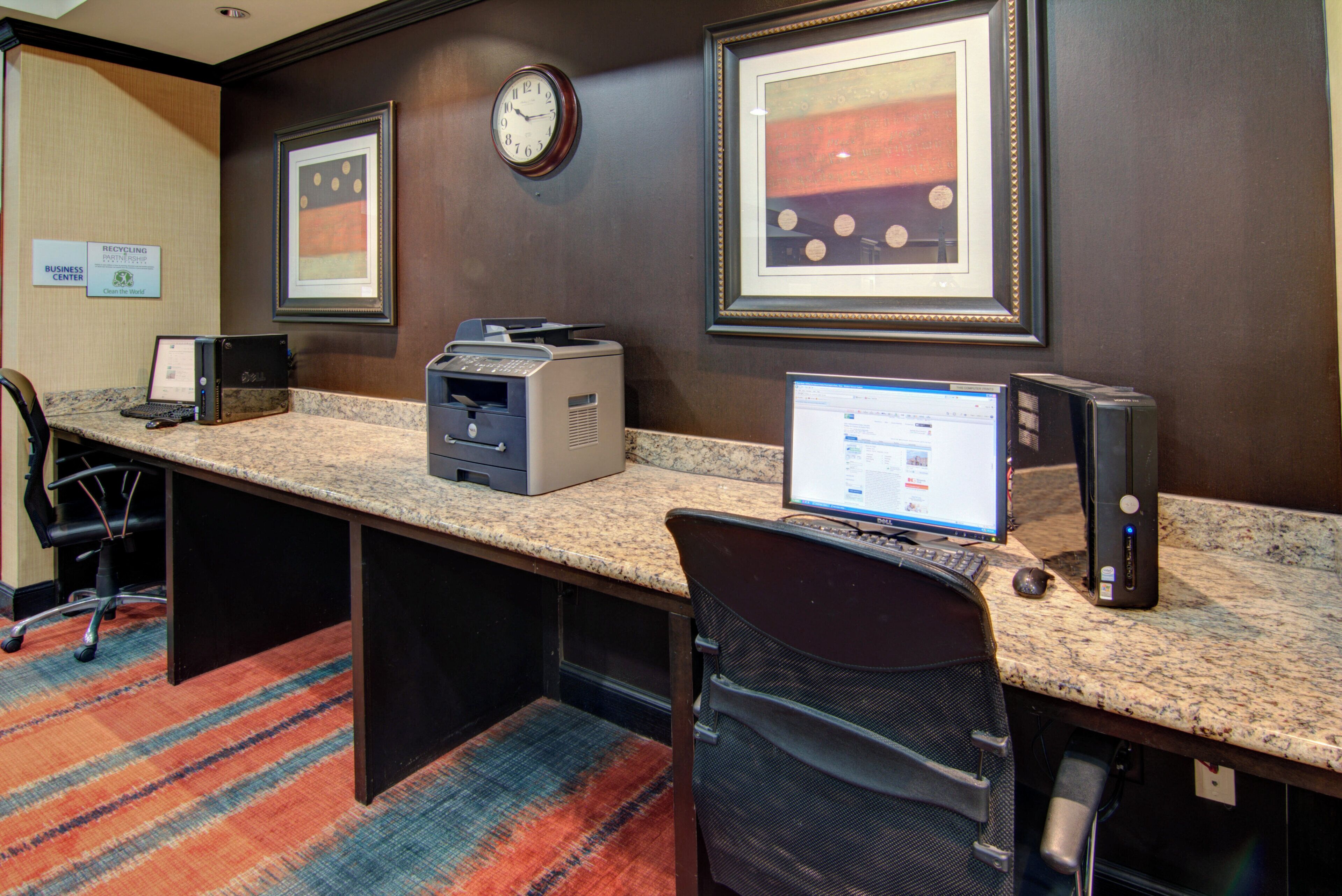 A hotel business center with a desktop computer, a printer, and a clock on the wall.