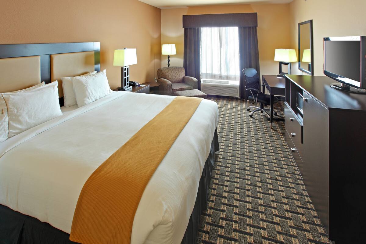 Ein Hotelzimmer mit einem großen Bett, einem Schreibtisch mit Stuhl, einem Fernseher und einem Fenster mit Vorhängen.