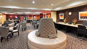A hotel lobby with a central seating area, multiple tables, and chairs, a reception desk, and a clock on the wall.
