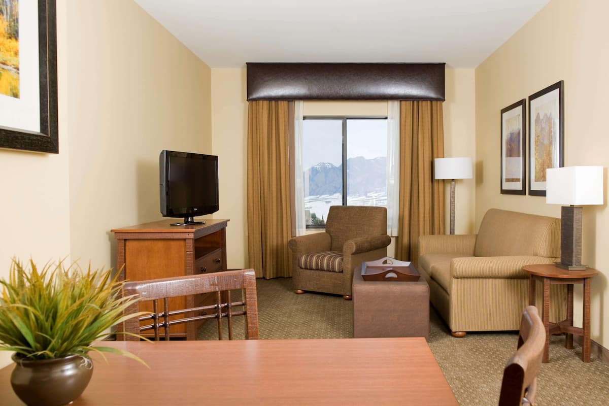A hotel room with a wooden desk, a television, a sofa, a chair, a small table, and a lamp.