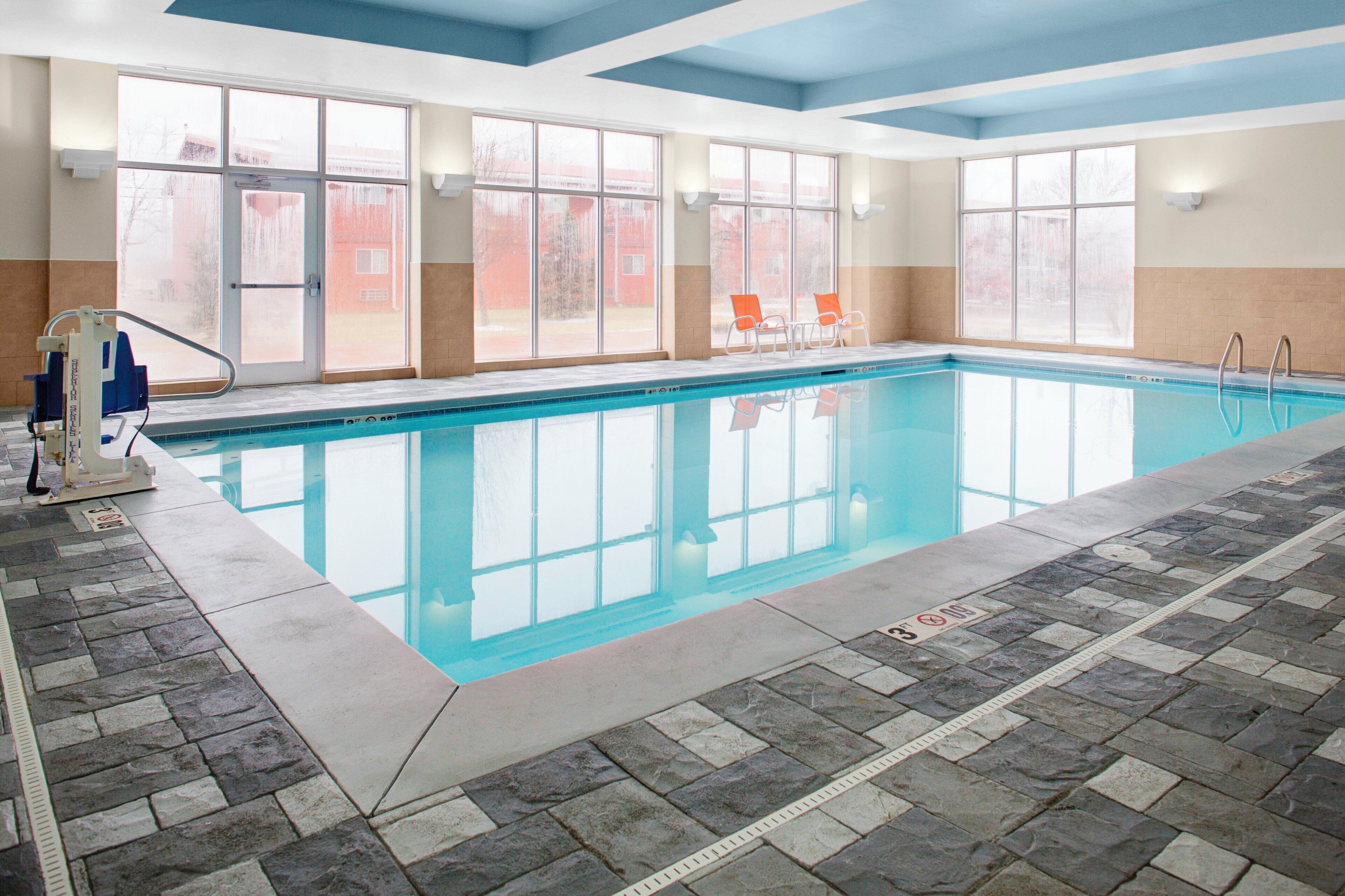 An indoor swimming pool with clear glass walls, a tiled floor, and a few lounge chairs.