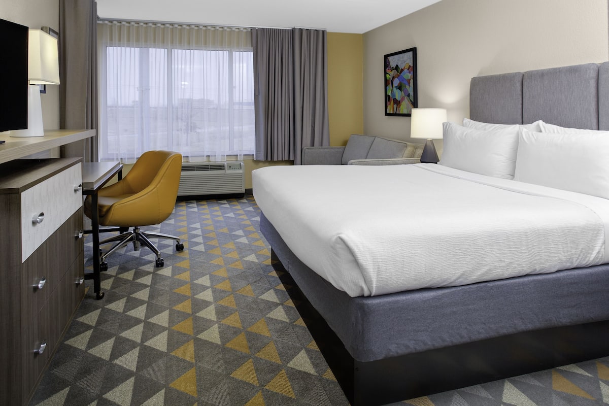 A hotel room with a large bed, a desk with a chair, a window with curtains, and a painting on the wall.