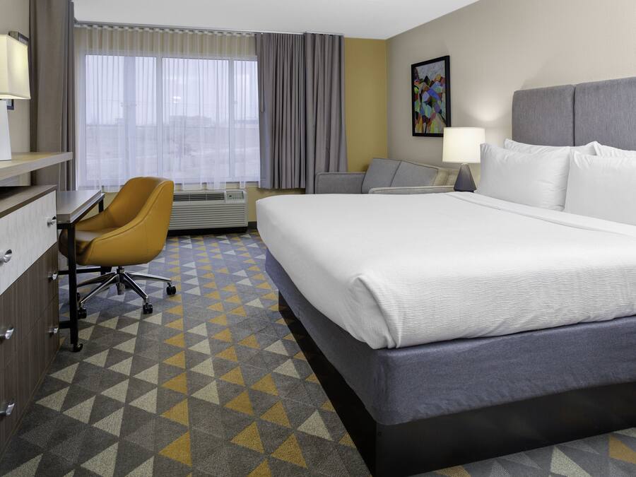 A hotel room with a large bed, a desk with a chair, a window with curtains, and a painting on the wall.