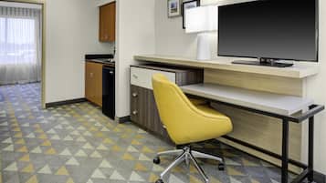 A hotel room with a desk, a yellow chair, a television, a kitchen area, and a patterned floor.