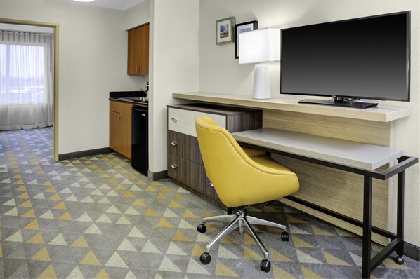 A hotel room with a desk, a yellow chair, a television, a kitchen area, and a patterned floor.