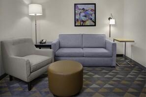 Une pièce avec un canapé gris, un fauteuil gris, un pouf rond, une petite table avec une lampe et un tableau abstrait encadré accroché au mur.