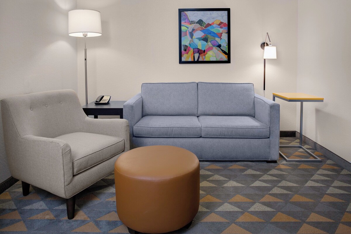 A room with a grey sofa, a brown ottoman, a small table, a lamp, a phone, and a colorful abstract painting on the wall.