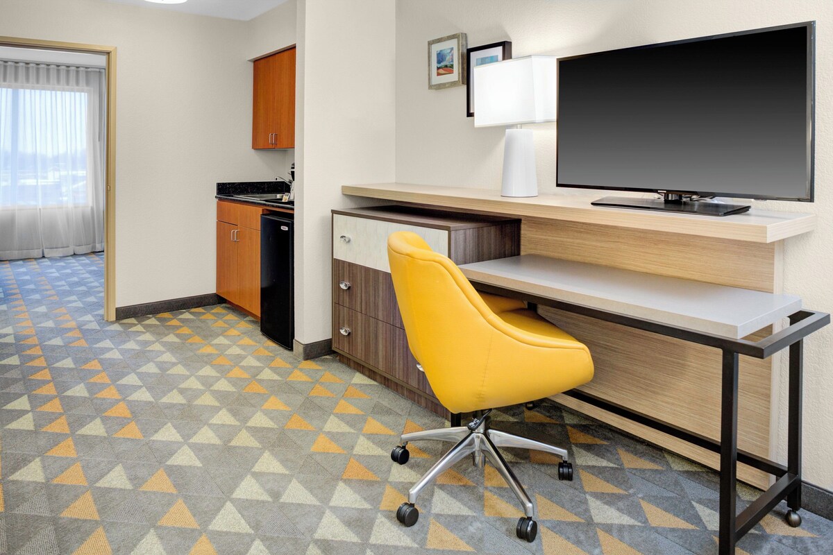 A hotel room with a desk, chair, TV, kitchen area, and patterned floor.