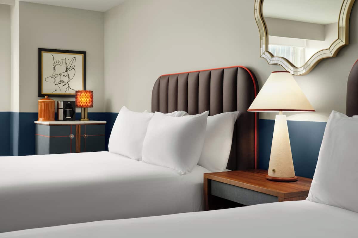 Hotellihuone, jossa on kaksi sänkyä, yöpöytä lampun kanssa, seinällä oleva kehystetty taideteos ja punaisella raidalla koristeltu kaappi.