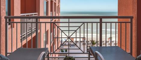 A balcony with two lounge chairs and a small table with a plant, overlooking a beach and ocean.