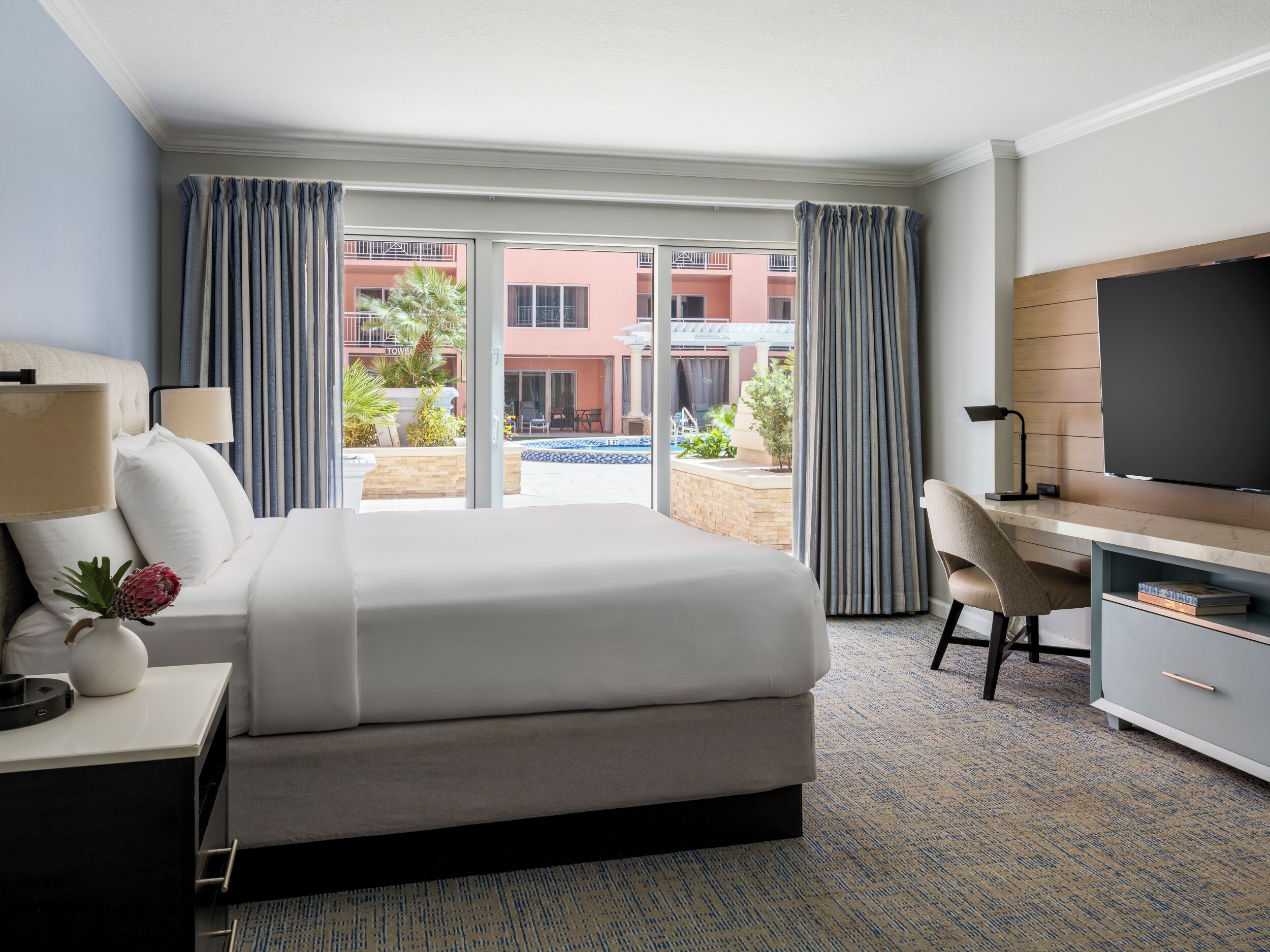 A hotel room with a bed, desk, TV, and a view of the outside building and pool.