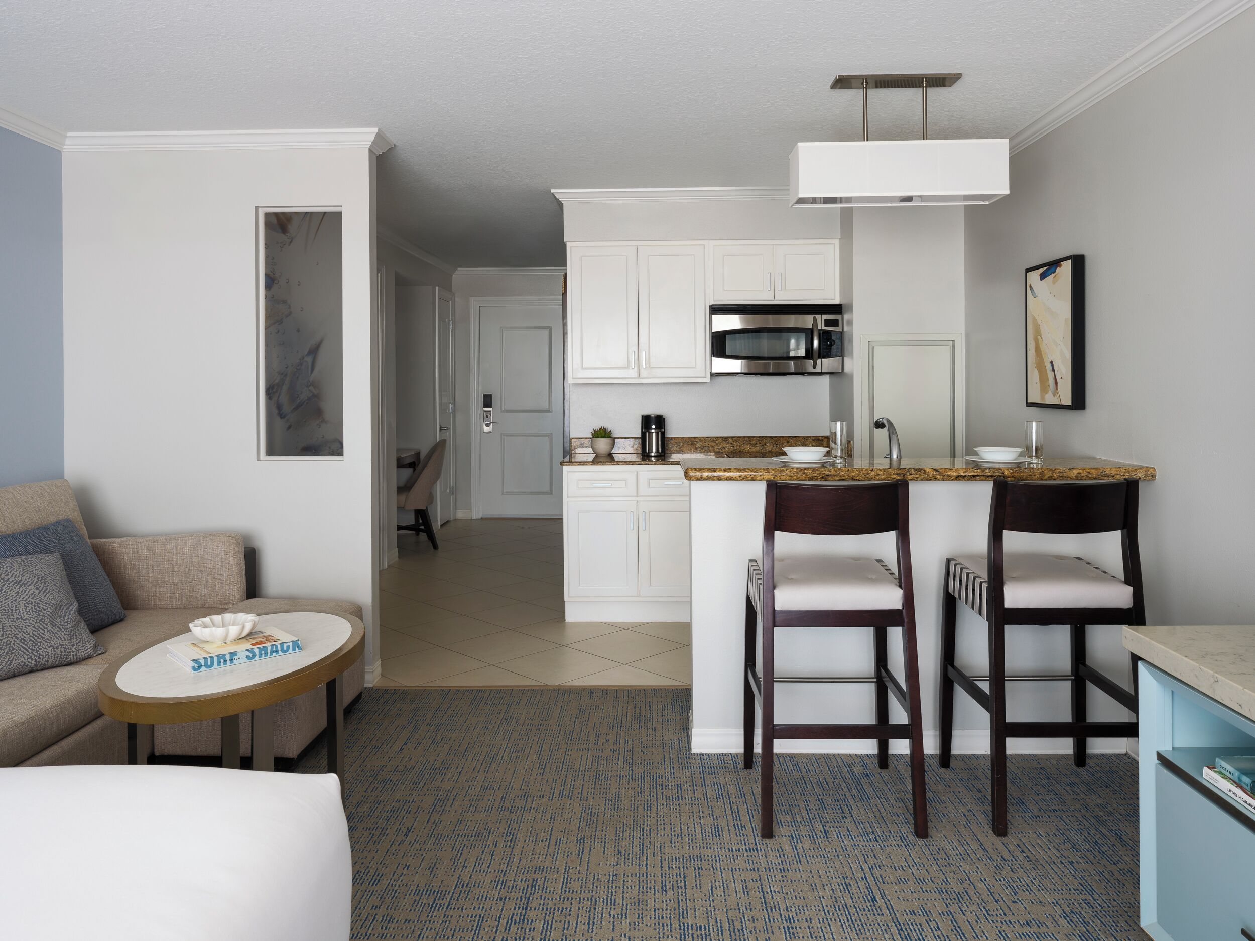 Una habitación de hotel moderna con kitchenette, un comedor con dos taburetes tipo bar, una pequeña mesa redonda con un libro y un sofá con un cojín.