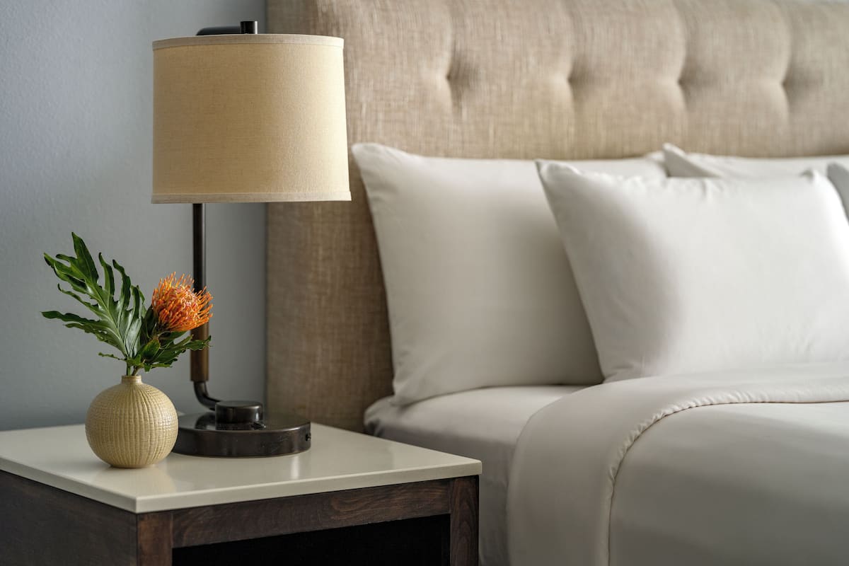 A neatly made bed with white linens, a bedside lamp, and a small vase with a plant on a bedside table.