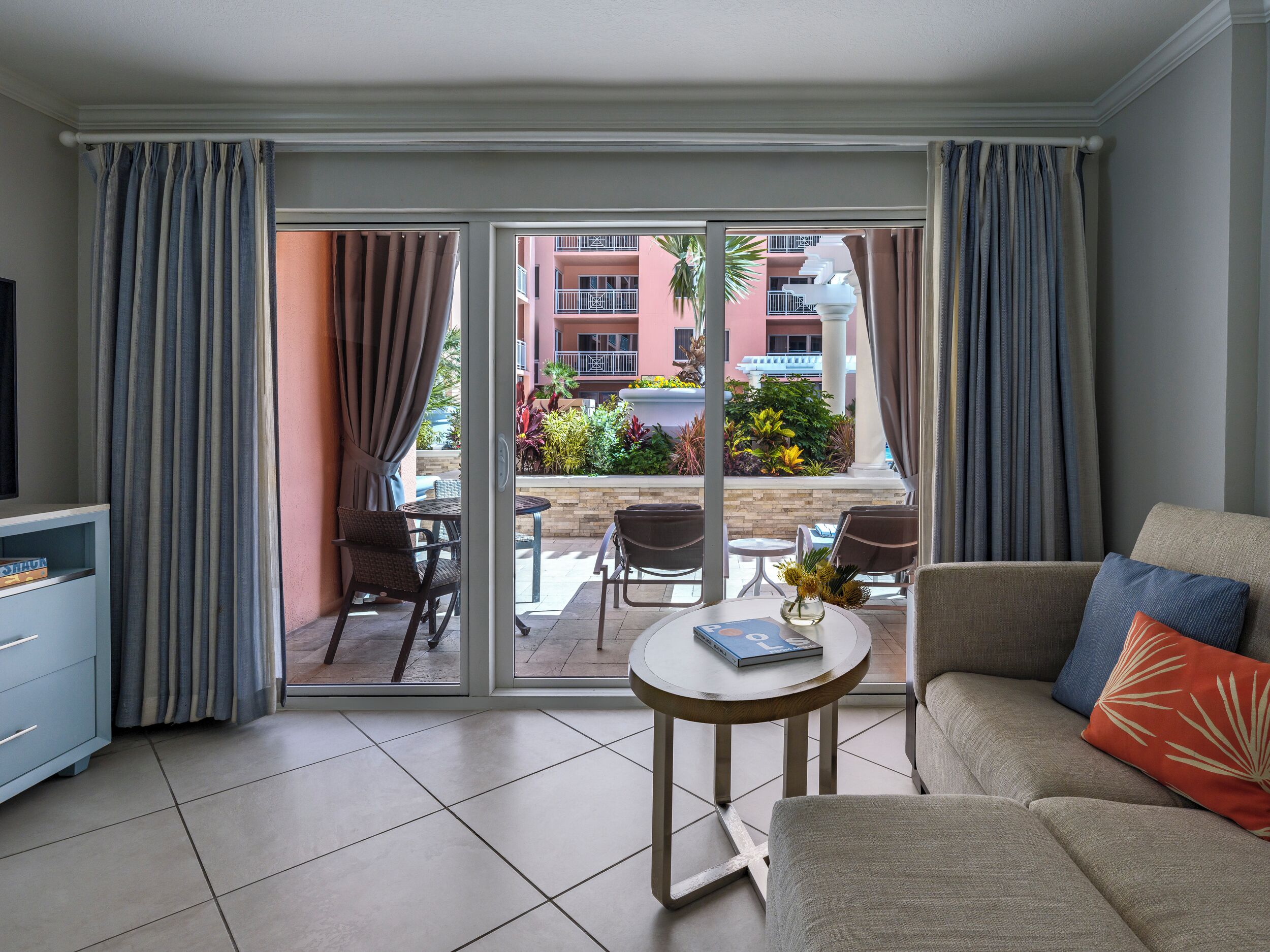 A hotel room with a sofa, a small table with a book, a TV, and a balcony with a view of a building and plants.