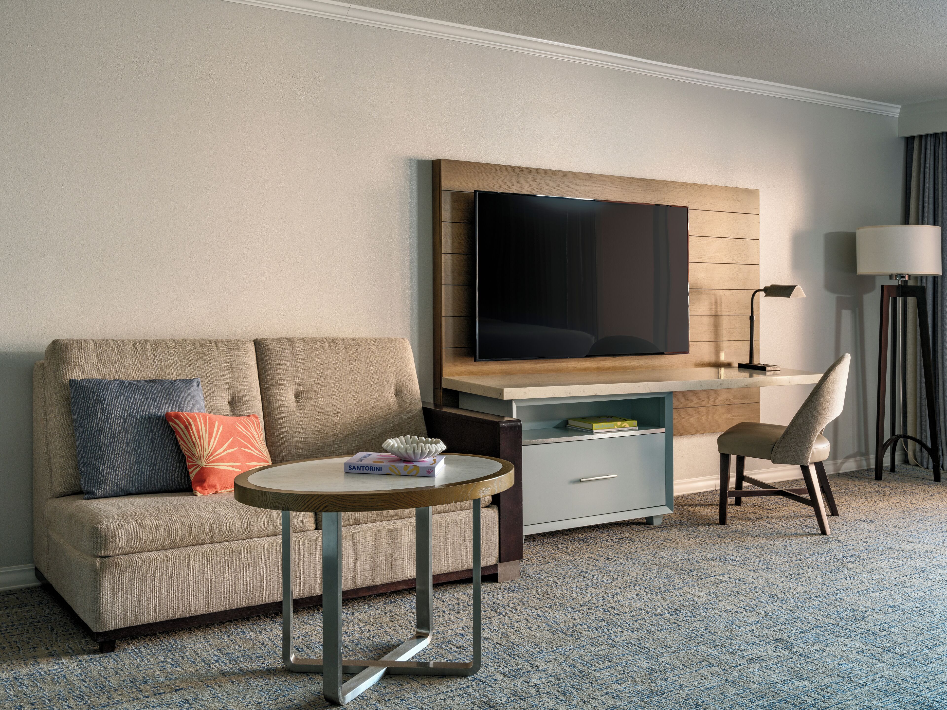 A hotel room with a sofa, a small round table, a TV, a desk, a chair, and a lamp.