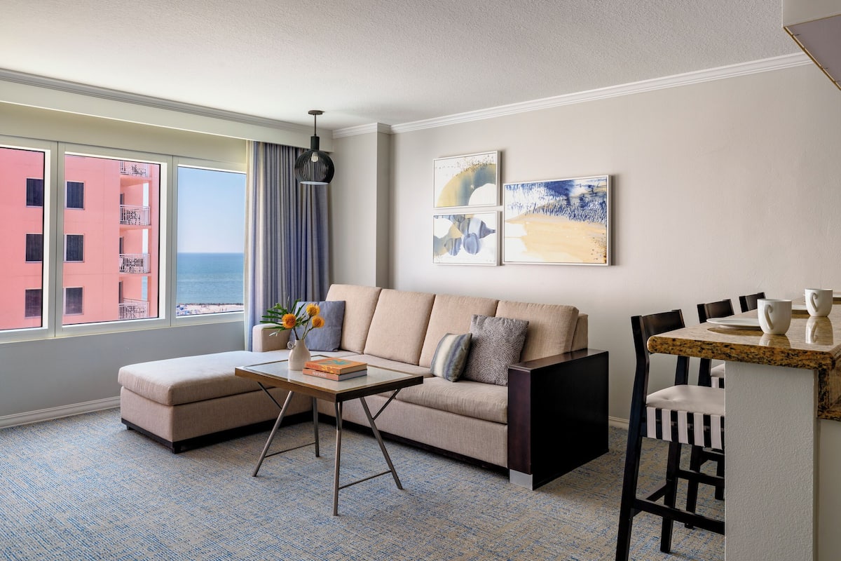 A modern living room with a sofa, coffee table, and dining area. A large window offers a view of the ocean and a pink building.