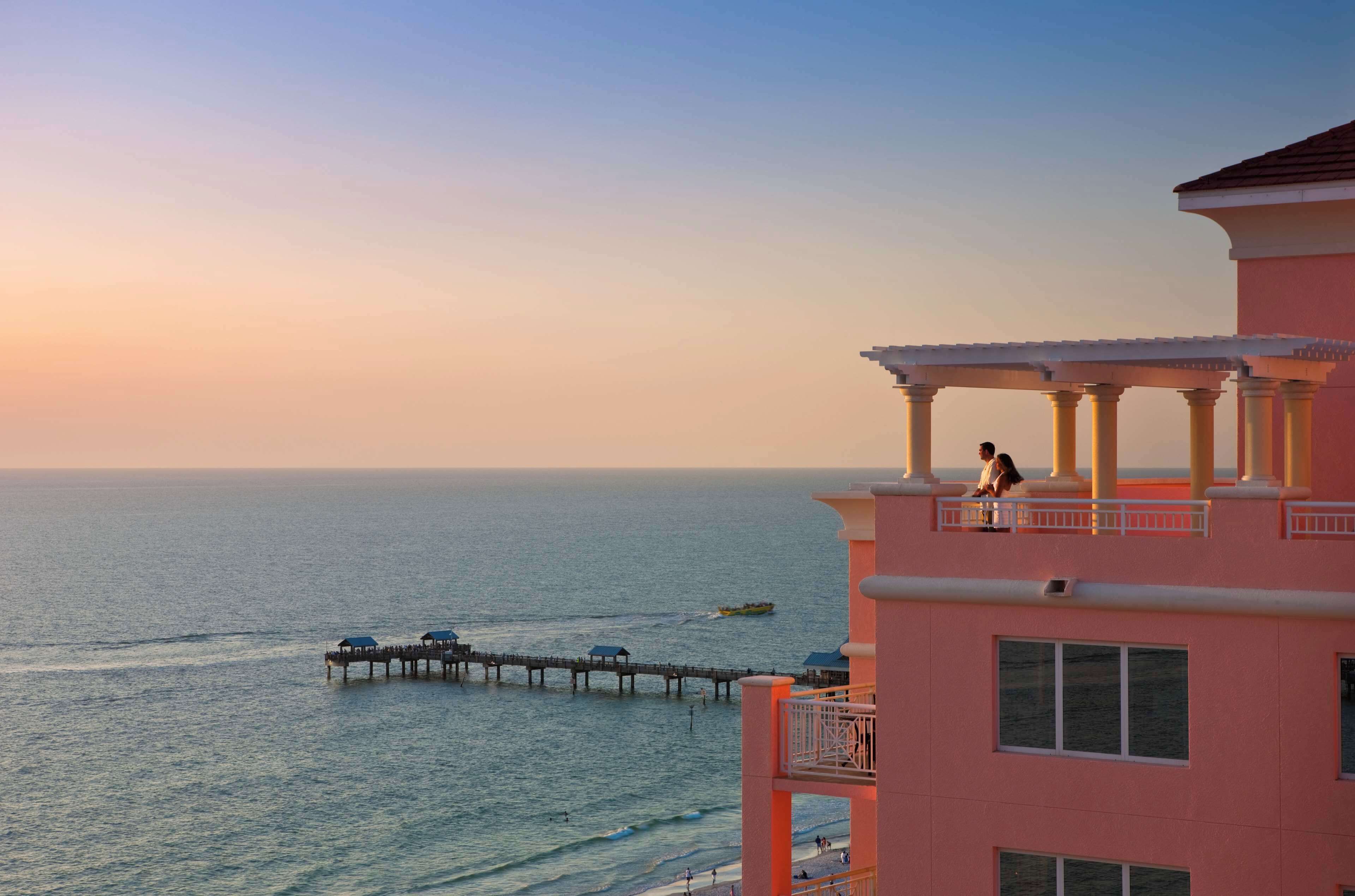 A pink building with a balcony overlooking the sea, a pier extending into the water, and a person standing on the balcony.