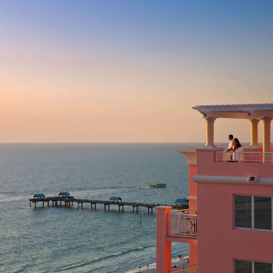 A pink building with a balcony overlooking the sea, a pier extending into the water, and a person standing on the balcony.