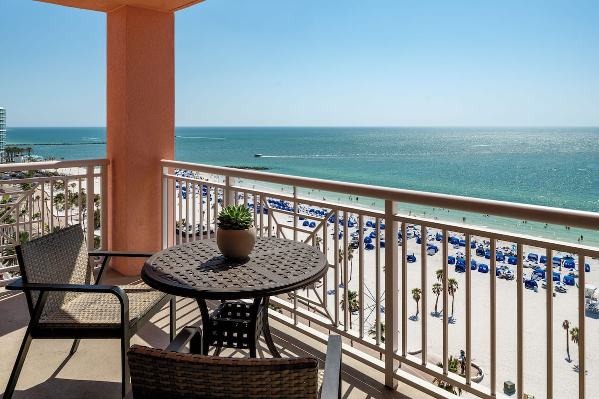 A balcony with a table and chairs, overlooking a beach with blue umbrellas and a clear view of the ocean.