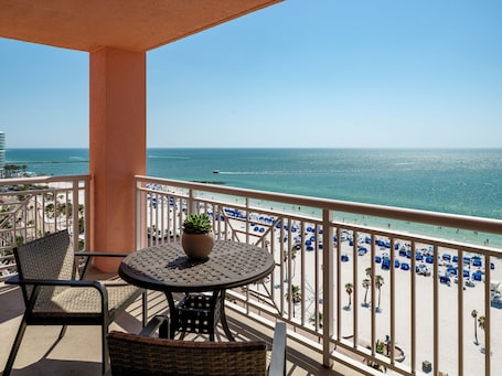 A balcony with a table and chairs, overlooking a beach with blue umbrellas and a clear view of the ocean.