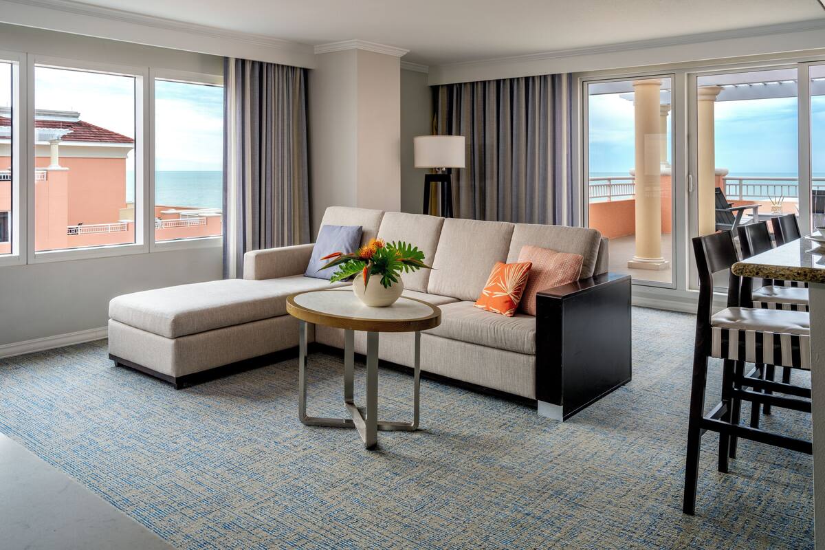 A modern hotel room with a sofa, a small round table, and a dining area with chairs.