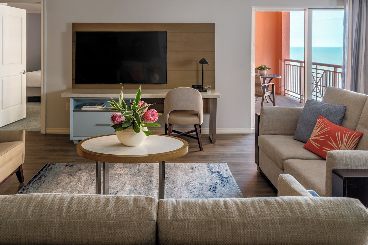 A modern living room with a flat-screen TV, a dining area, and a balcony with a view of the sea.