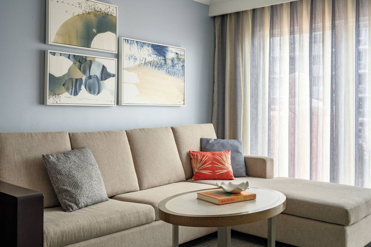 A modern living room with a beige sectional sofa, a round wooden coffee table, and a book titled "MALDIVES" on it. There are two framed artworks above the sofa and a window with sheer curtains to the right.