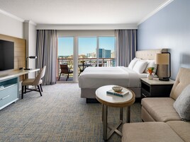 Habitación de hotel con cama, escritorio, televisión, balcón con vistas a la ciudad y un área de estar con sofá y lámpara.