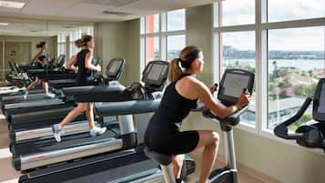 A gym with treadmills and an elliptical machine, large windows with a city view, and a mirror reflecting the activity.