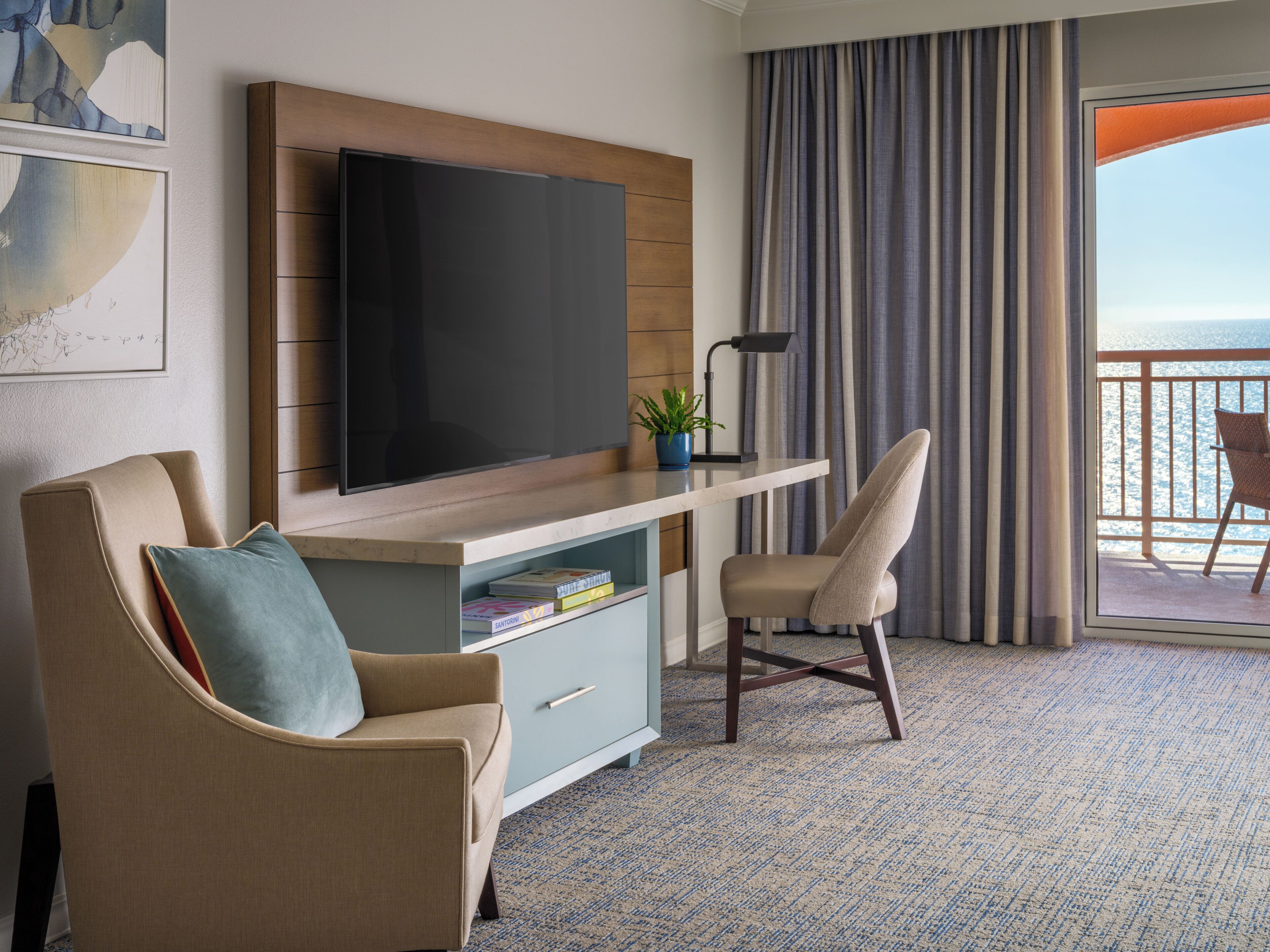 Une chambre d’hôtel équipée d’une télévision à écran plat, d’un bureau avec une lampe, d’une chaise, d’une bibliothèque et offrant une vue sur la mer à travers les rideaux.
