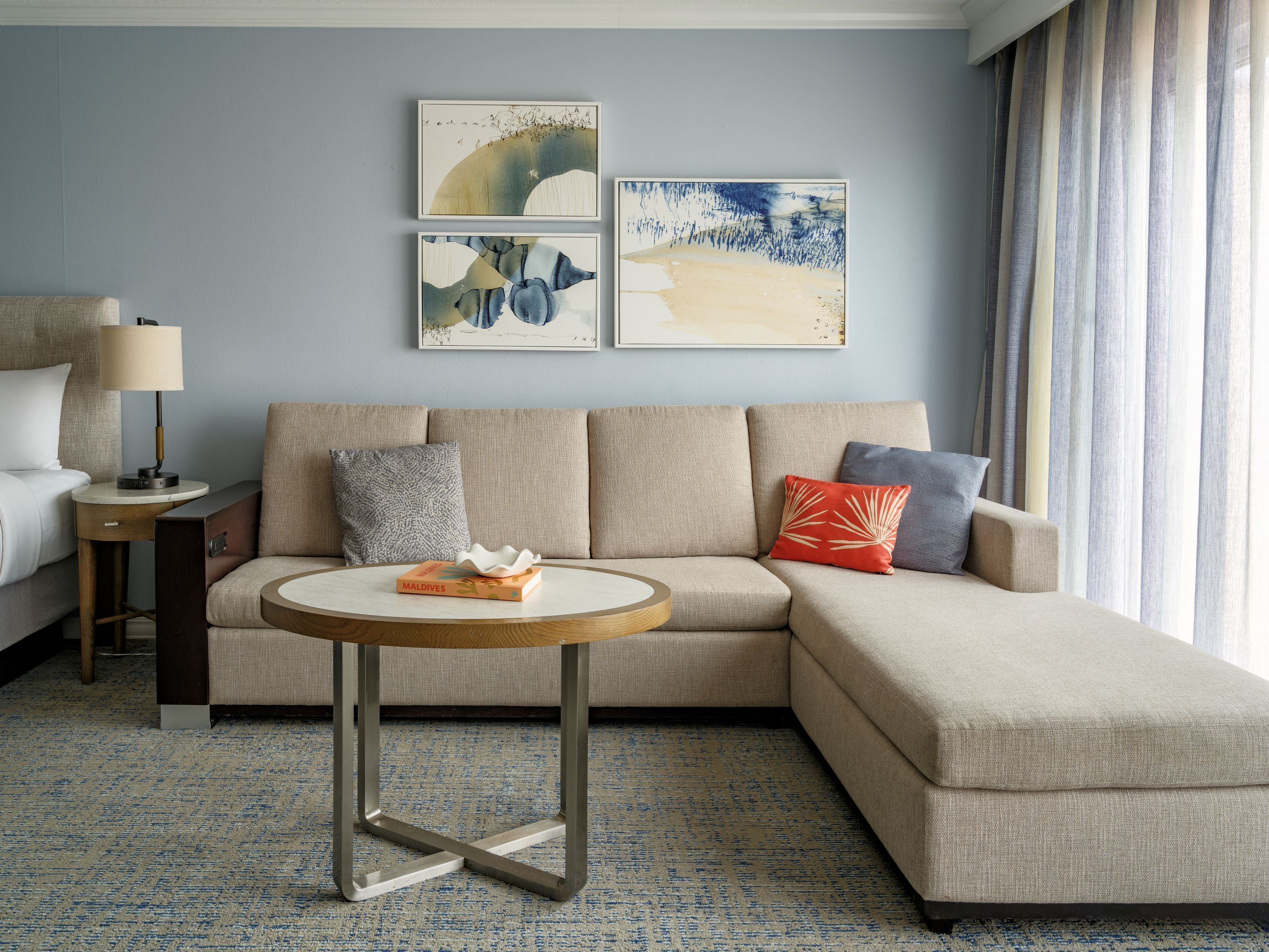 Et moderne hotelværelse med en beige sofa, et rundt sofabord og en seng med hvid dyne. Der er tre billeder hængende på væggen, en lampe på et sengebord og et vindue med gardiner.
