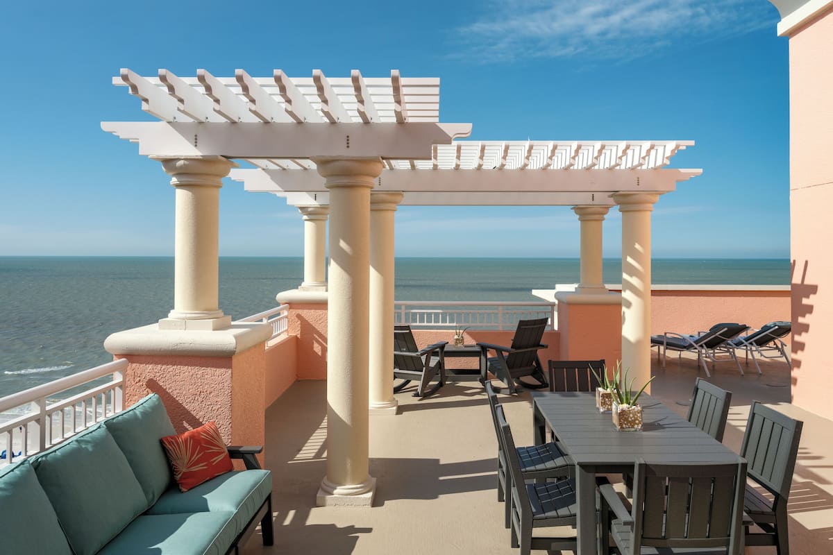 A balcony with a view of the ocean, featuring a pergola, seating area, and a table with chairs.