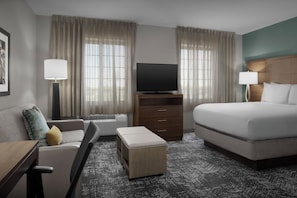 Habitación de hotel con cama, televisor en un mueble bajo, sofá, escritorio y ventana con cortinas.