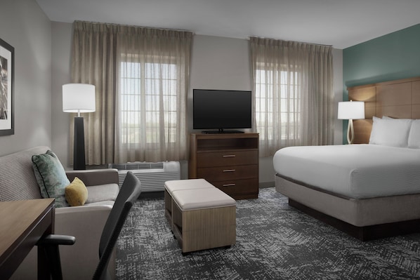 A hotel room with a bed, a TV on a stand, a sofa, a desk, and a window with curtains.